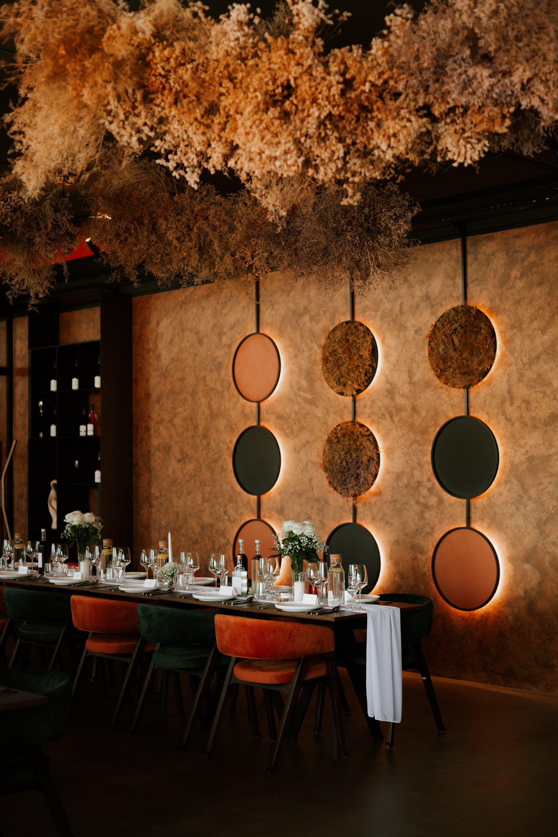 Elegantná svadobná hostina — dlhá tabuľa so zamatovými stoličkami v odtieňoch rdze a lesnej zelene, inštalácia zo sušených kvetov a podsvietené dekoračné kruhy na stene