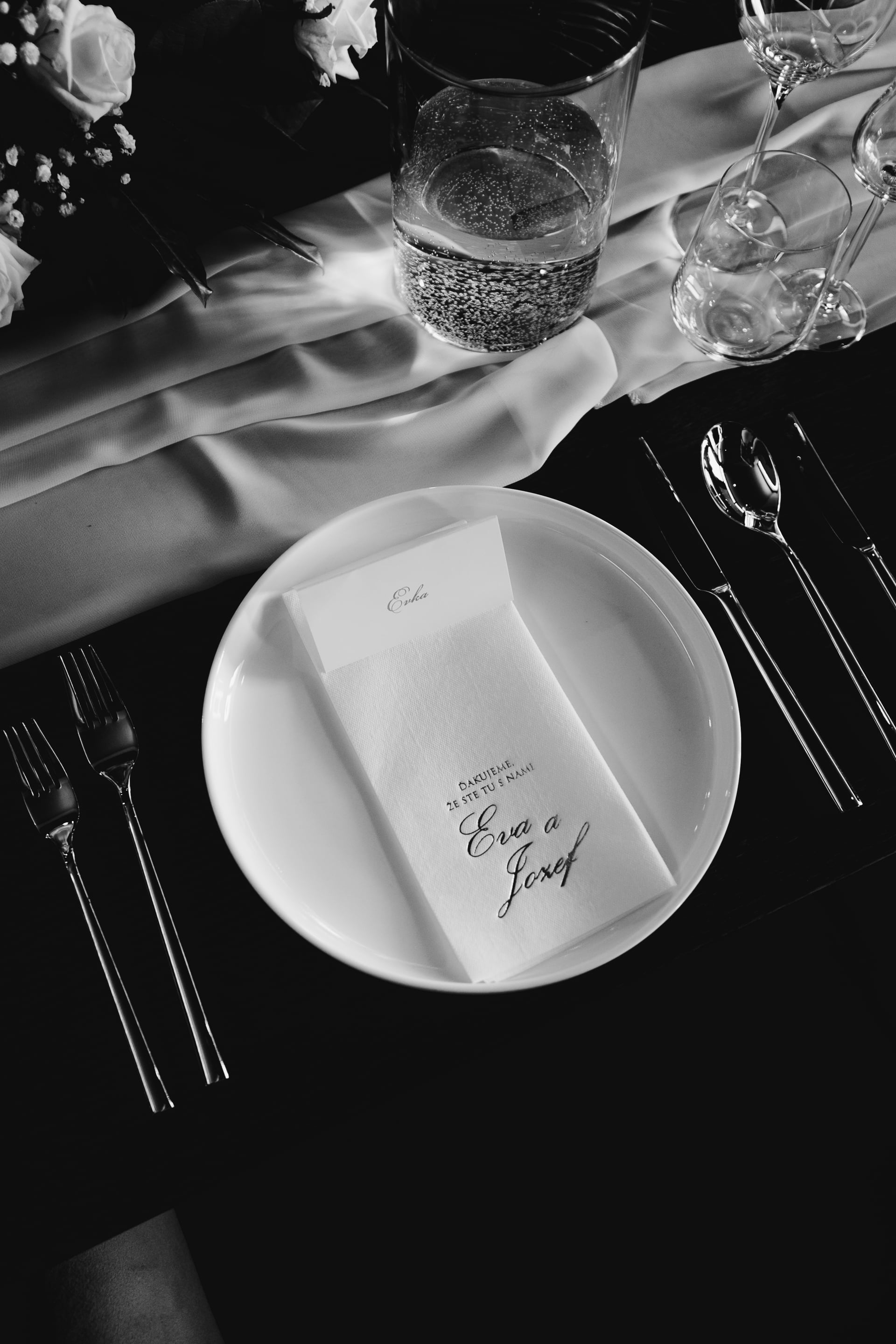 Čiernobiely pohľad zhora na svadobné prestieranie — biely tanier s menovkou a menu, strieborné príbory, pohár s perlivou vodou a biele ruže na hodvábnom behúni