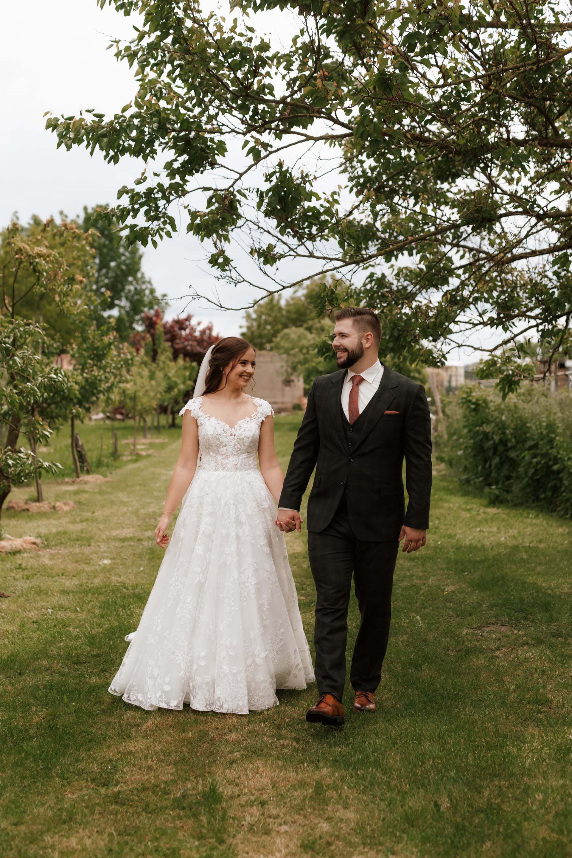 Nevesta v čipkovaných šatách so závojom a ženích v tmavom obleku s vínovou kravatou kráčajú rukou v ruke po tráve v ovocnom sade — vzájomný pohľad s úsmevom