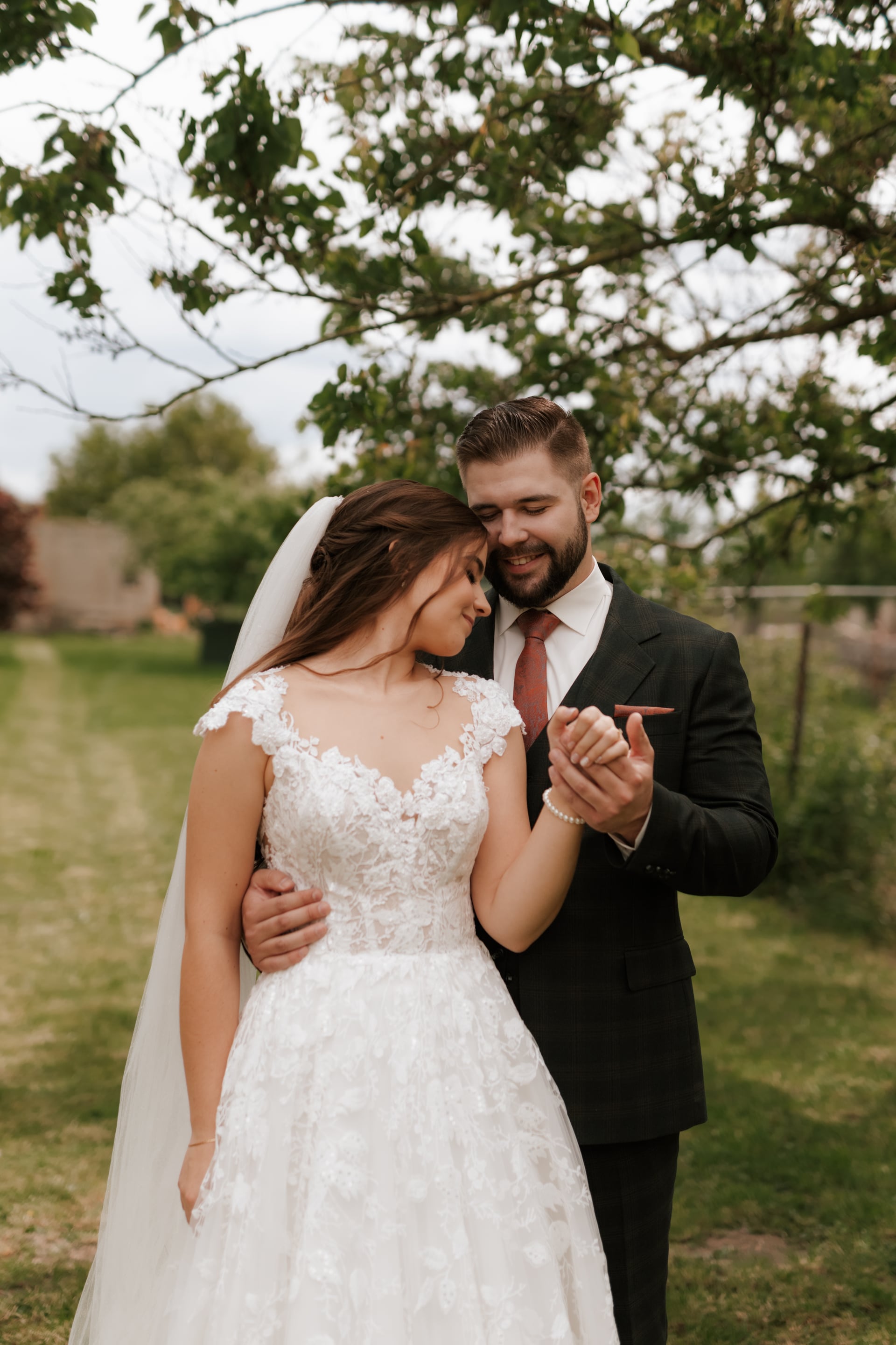 Svadobný portrét v záhrade — nevesta v čipkovaných šatách so závojom a ženích v tmavom obleku s vínovou kravatou si prezerajú prsteň na jej ruke pod konármi ovocného stromu