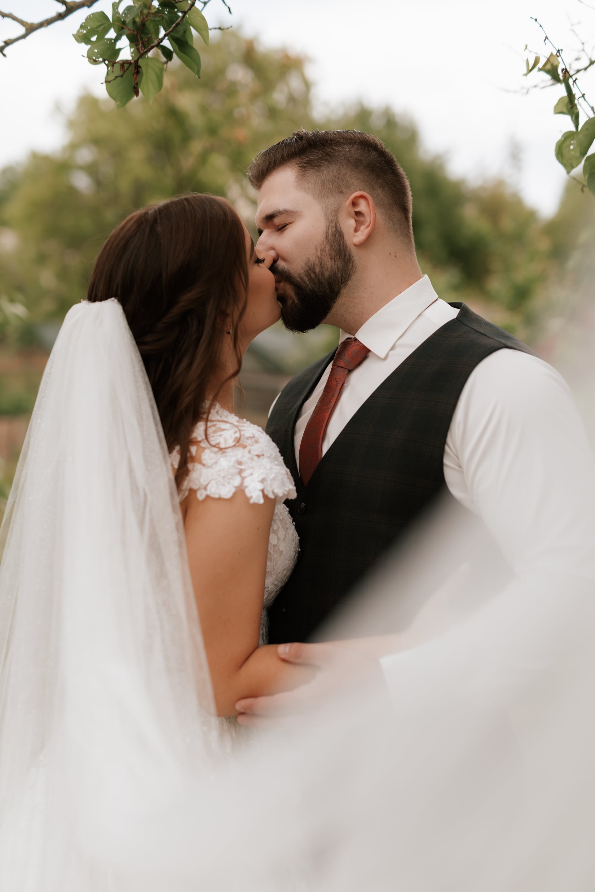 Intímny bozk nevesty v čipkovaných šatách a ženícha v károvanom vestíku s vínovou kravatou — vznášajúci sa závoj v záhrade