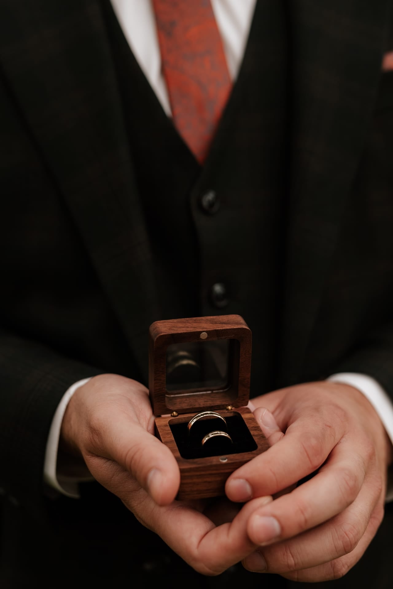 Detail ženíchových rúk s otvoreným dreveným puzdrom s dvoma zlatými obrúčkami — čierny trojdielny oblek s bielou košeľou a vínovou vzorovanou kravatou