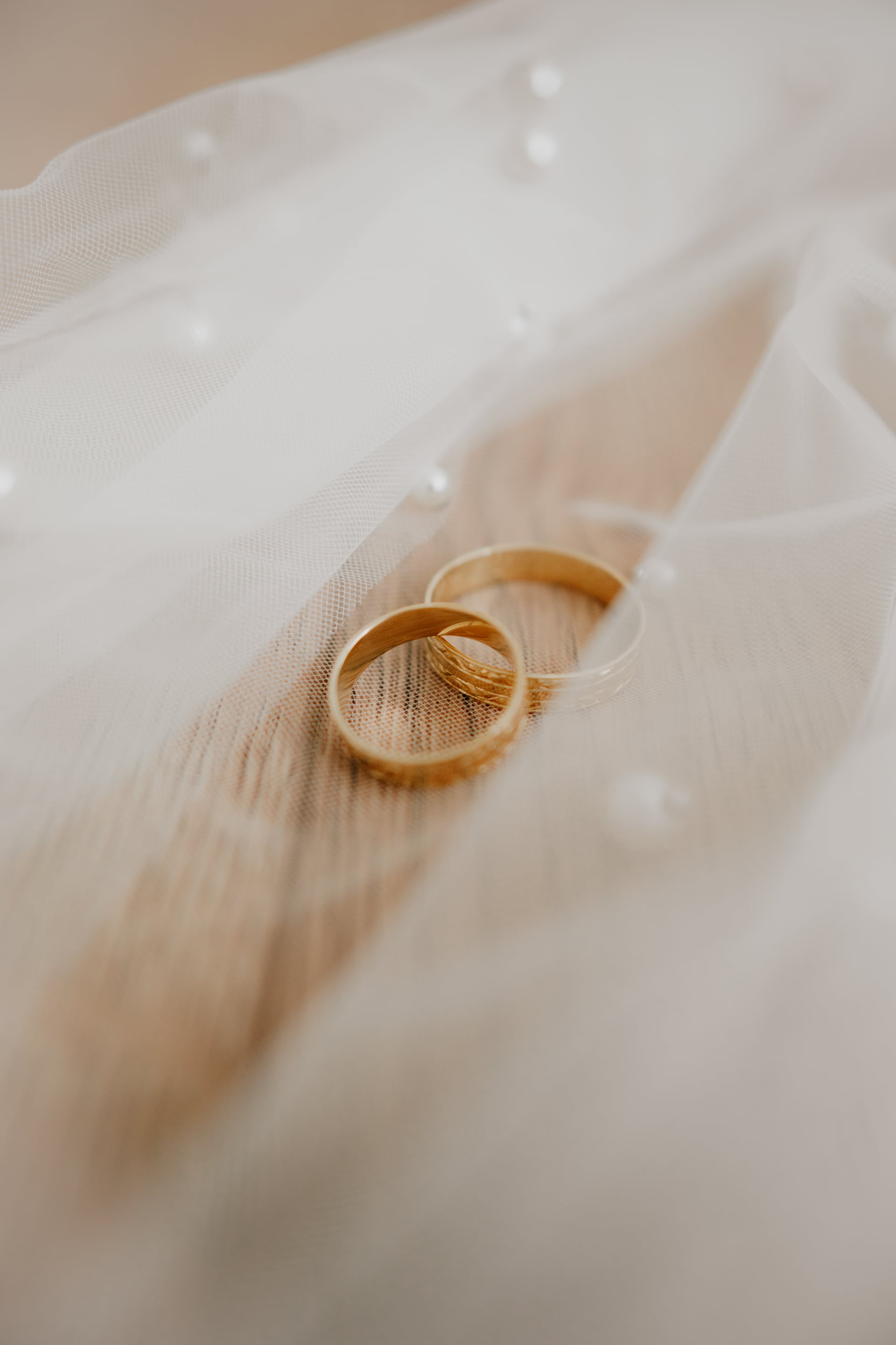 Detail dvoch zlatých svadobných obrúčok položených na bielom tylovom závoji s drobnými perličkami v jemnom dennom svetle