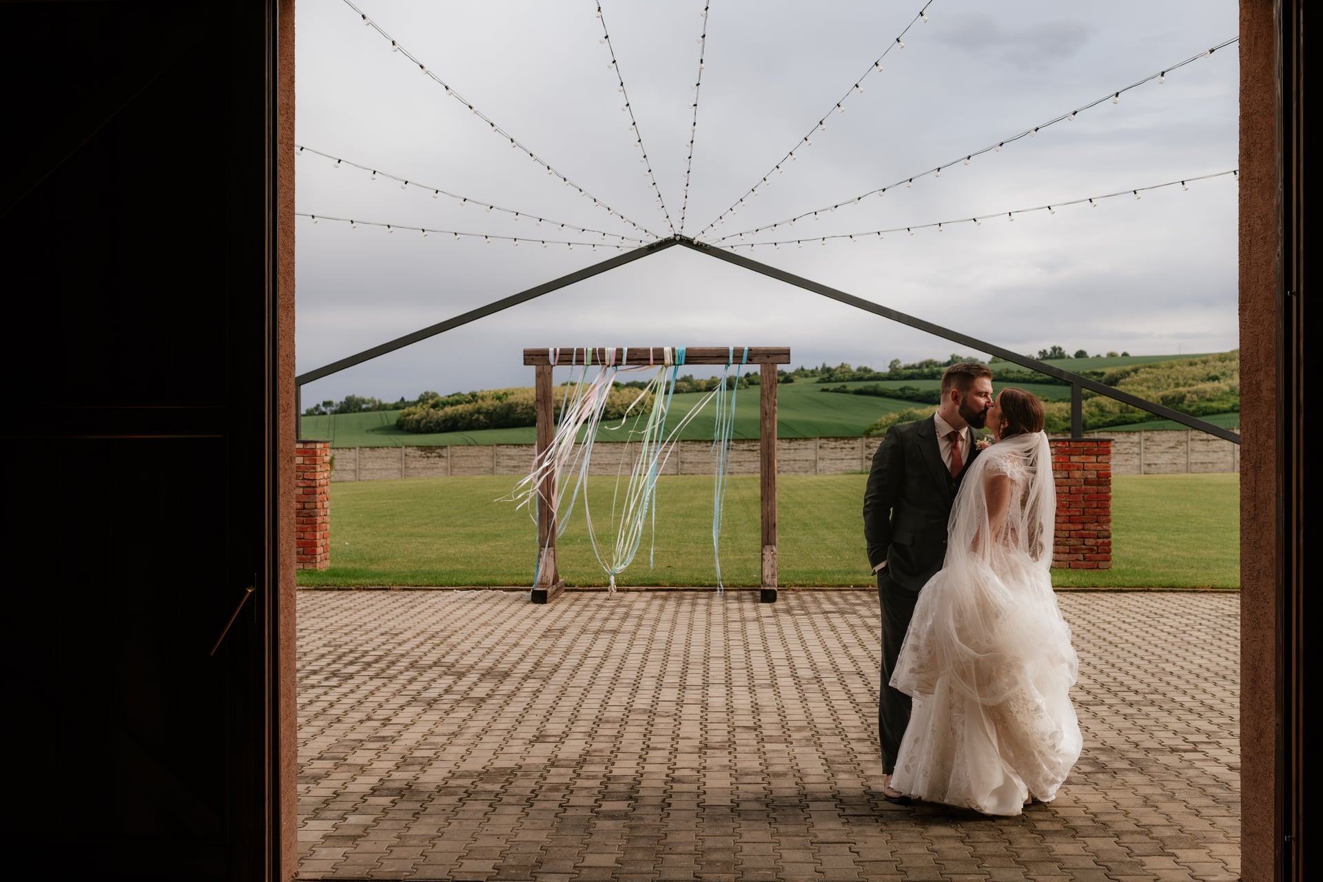 Nevesta so závojom a ženích v šedom obleku stoja pod drevenou pergolou s vlajúcimi stuhami — pohľad cez otvorený vchod stodoly na rozvlnenú krajinu so svetelnými lankami nad hlavou