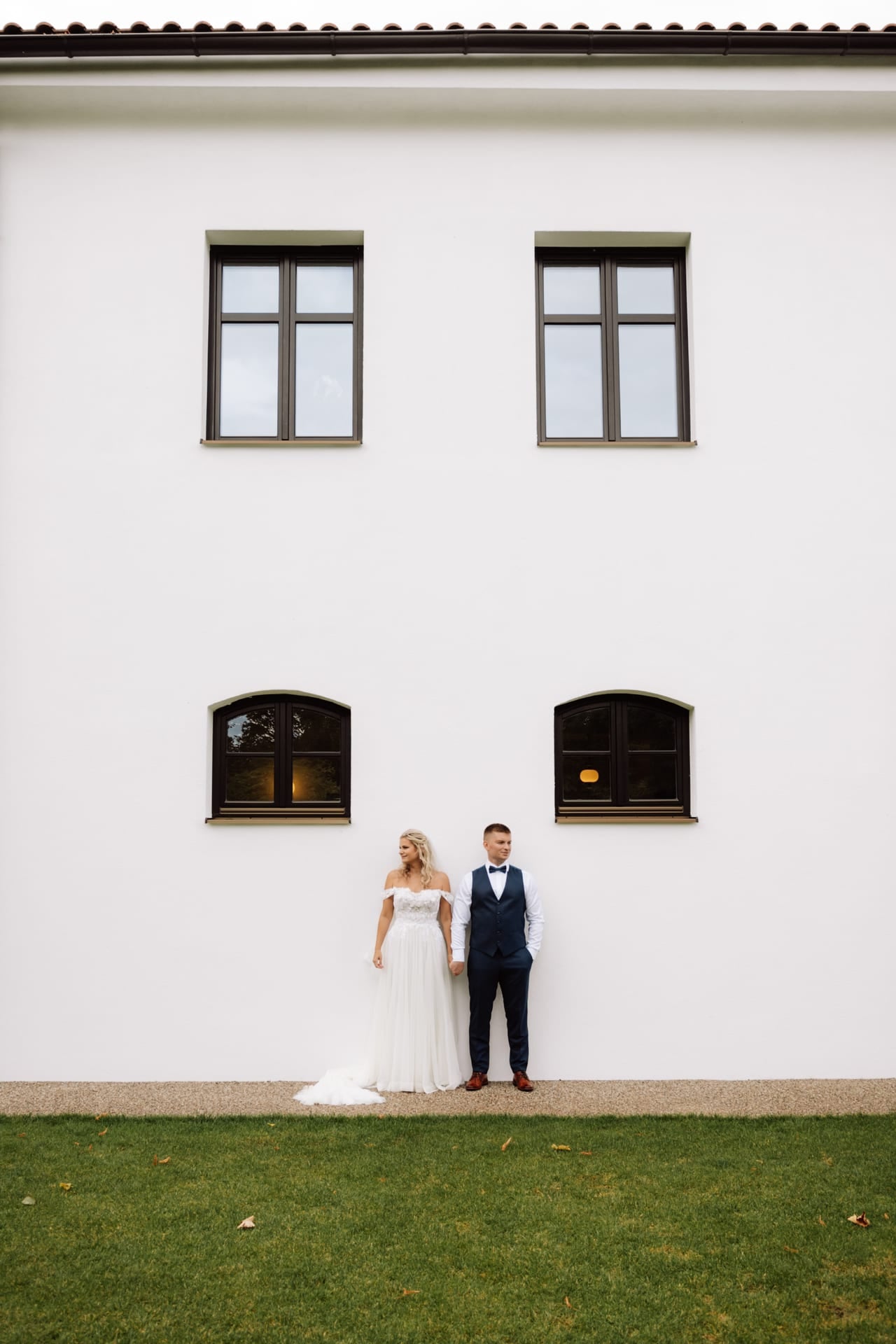 Nevesta v tylových šatách a ženích v modrom obleku držia sa za ruky pri bielej omietnutej stene kaštieľa s dreveným oknami a červenou strechou — široký záber s trávnatou plochou