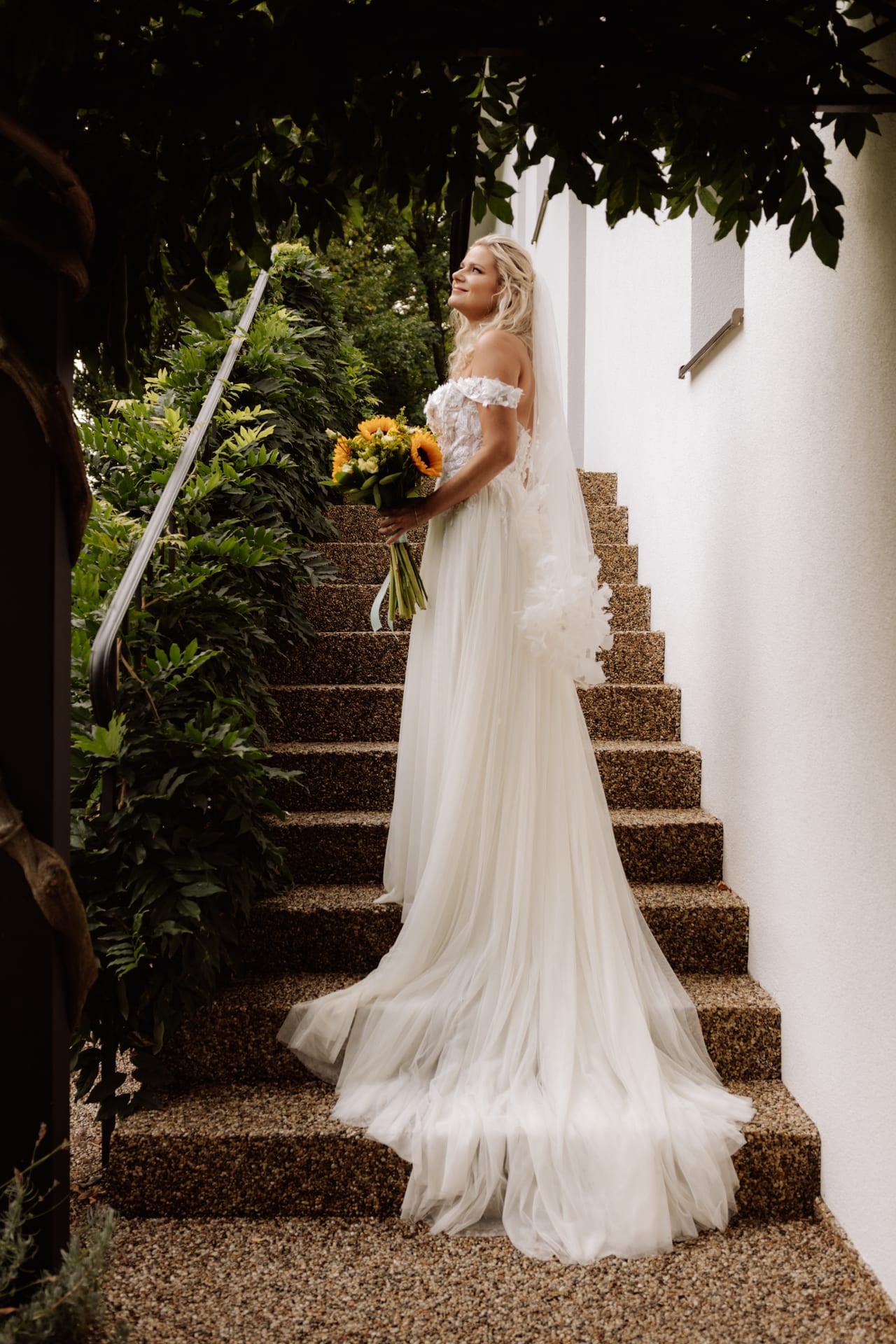 Nevesta v tylových šatách s čipkovaným korzetom a dlhým závojom stojí na kamenných schodoch so slnečnicovou kyticou — úsmev s pohľadom nahor