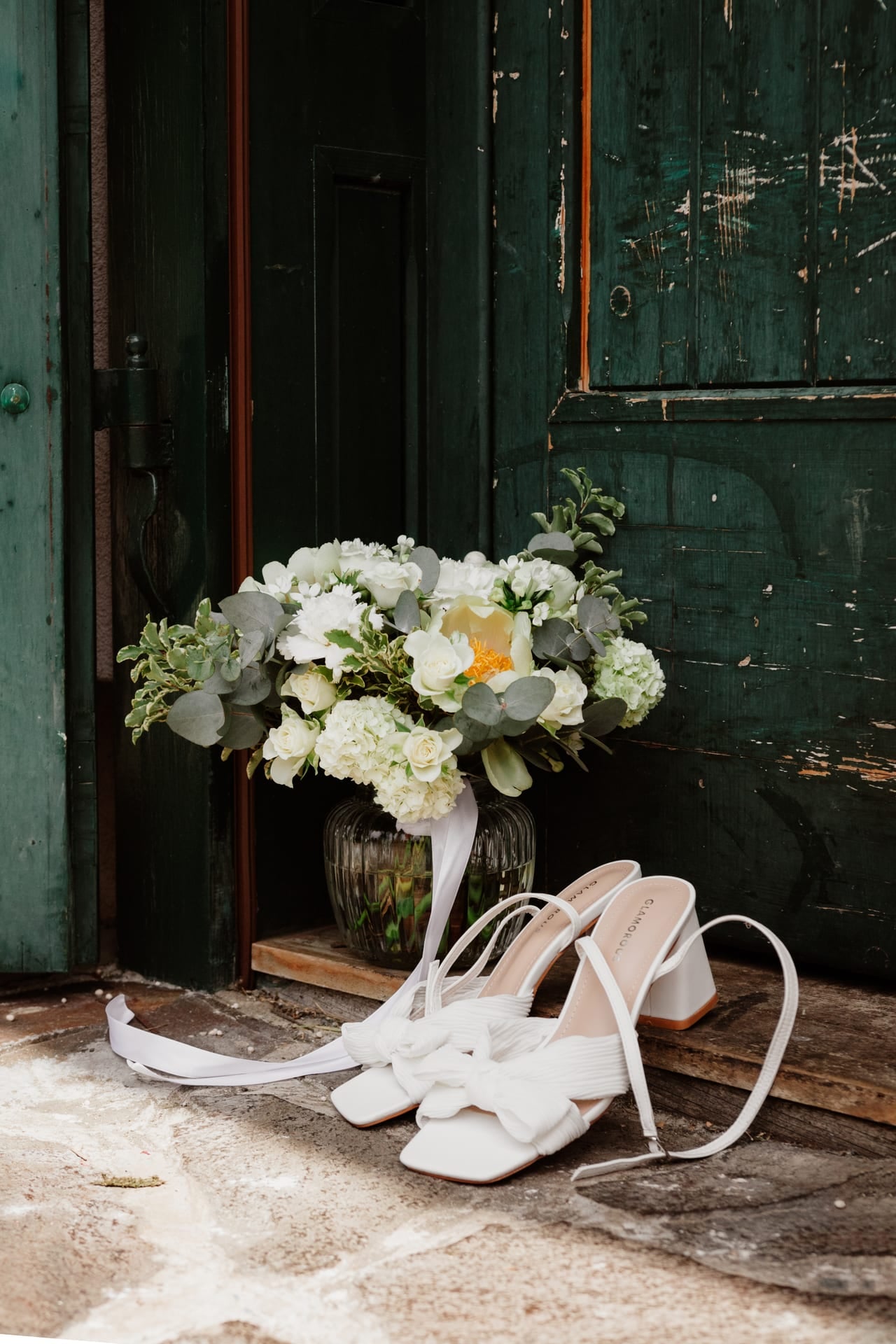 Detail svadobných detailov — biele remienkové topánky s mašľou a kytica z bielych hortenzií, ruží a eukalyptu v sklenej váze pred starými zelenými dverami