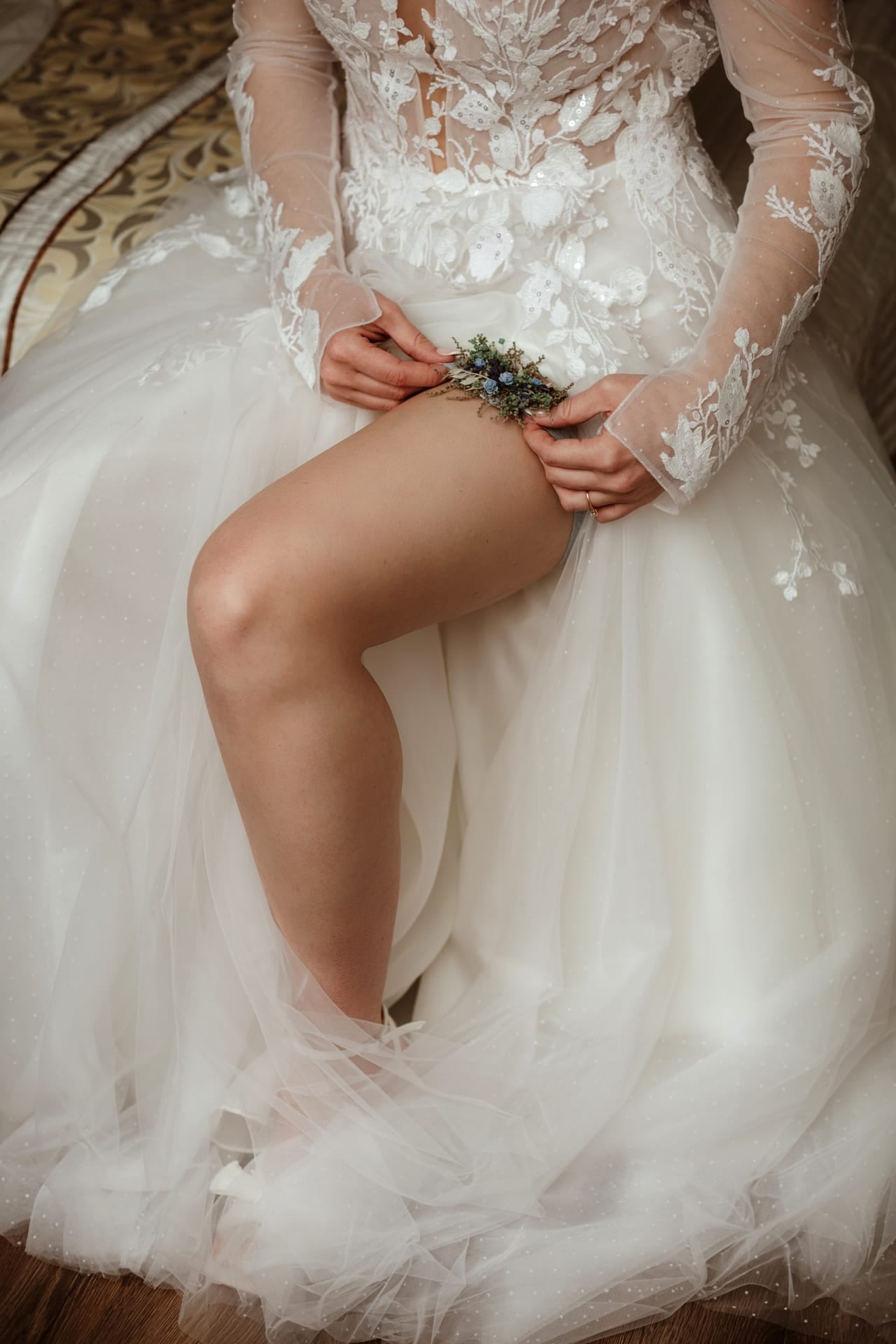 Detail svadobného podväzku s malými modrými sušenými kvetmi na nevestinom stehne — čipkové šaty s dlhými tylovými rukávmi so zdobením