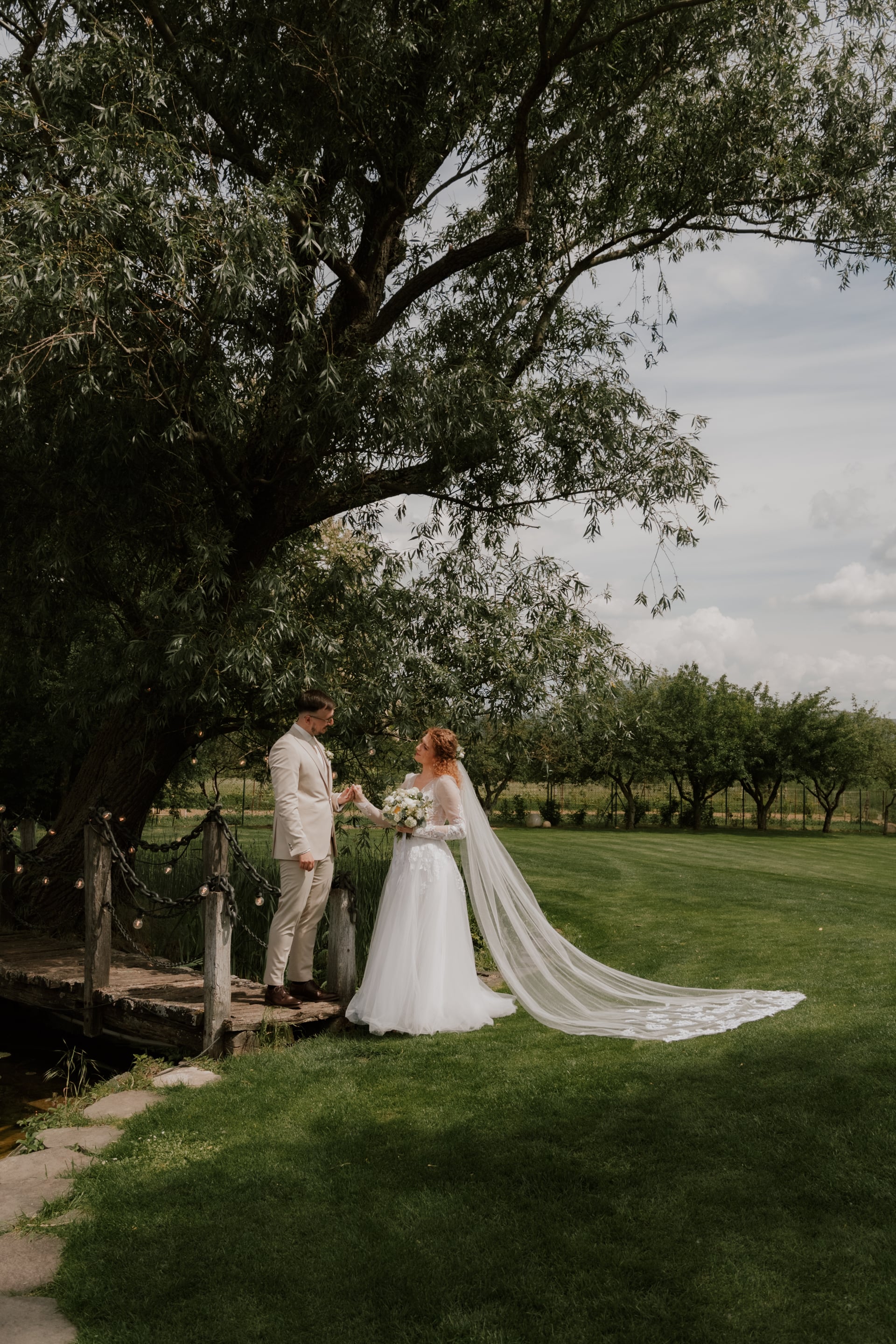 Nevesta s dlhými čipkovými rukávmi a dlhou vlečkou závoja a ženích v béžovom obleku stoja na drevenom mostíku pod vŕbou — svetelné lanká na zábradlí a trávnatá záhrada