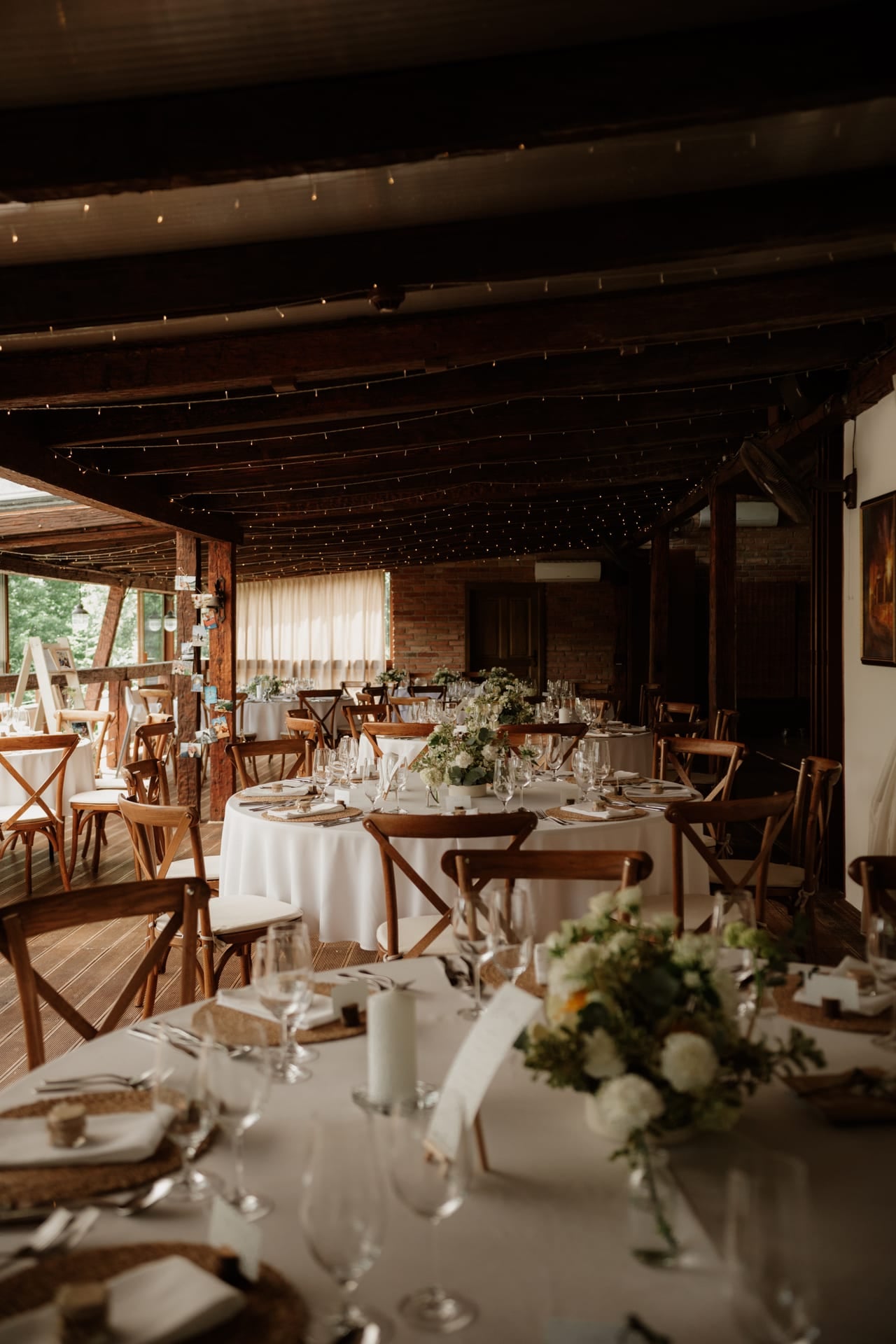Interiér rustikálnej svadobnej sály pred hostinou — okrúhle stoly s bielymi obrusmi a kvetinovými stredmi, drevené crossback stoličky a svetielka na drevených trámoch