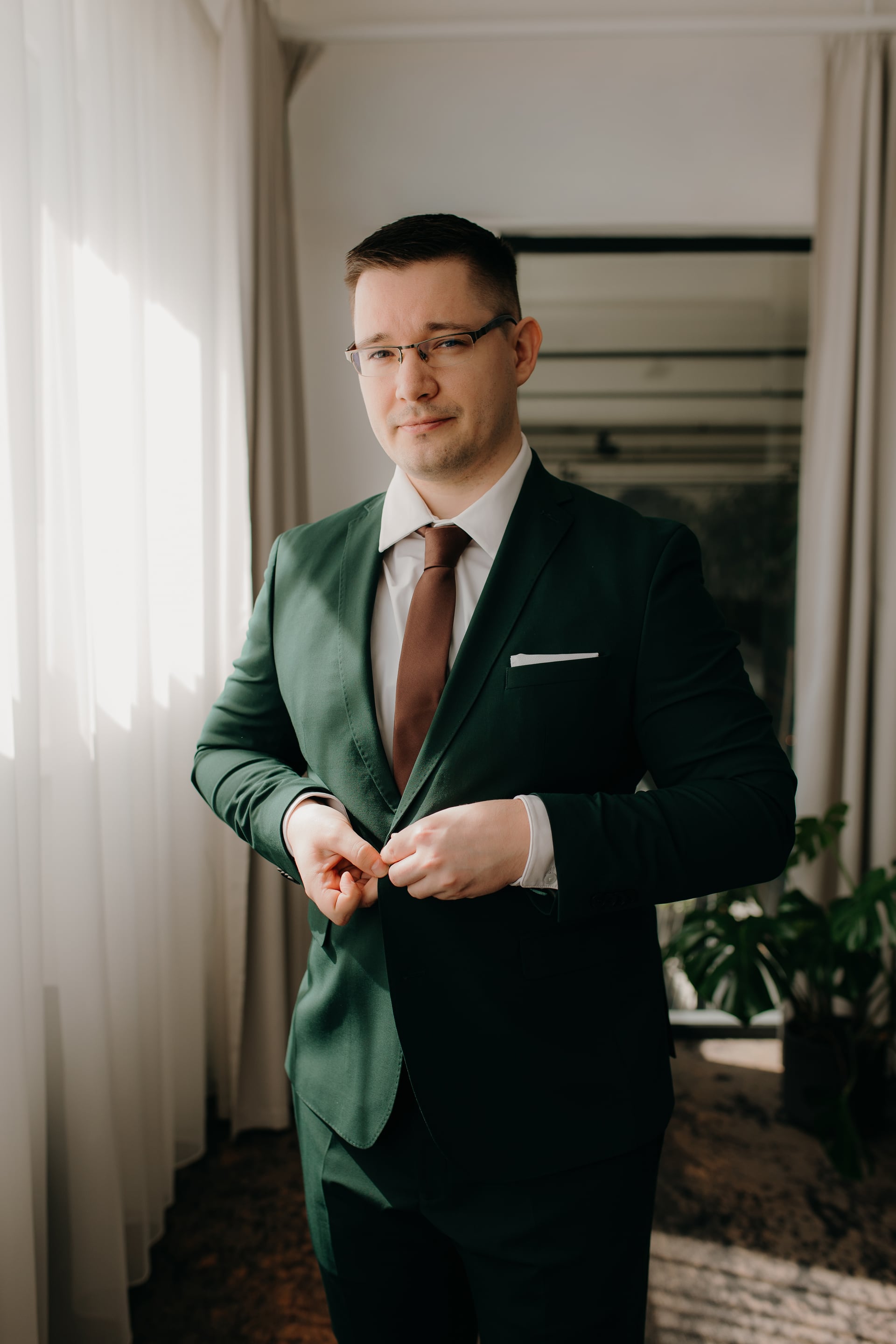 Ženích si zapína smaragdovozelené sako pri okne — hnedá kravata, biela vreckovka a okuliare v prírodnom svetle