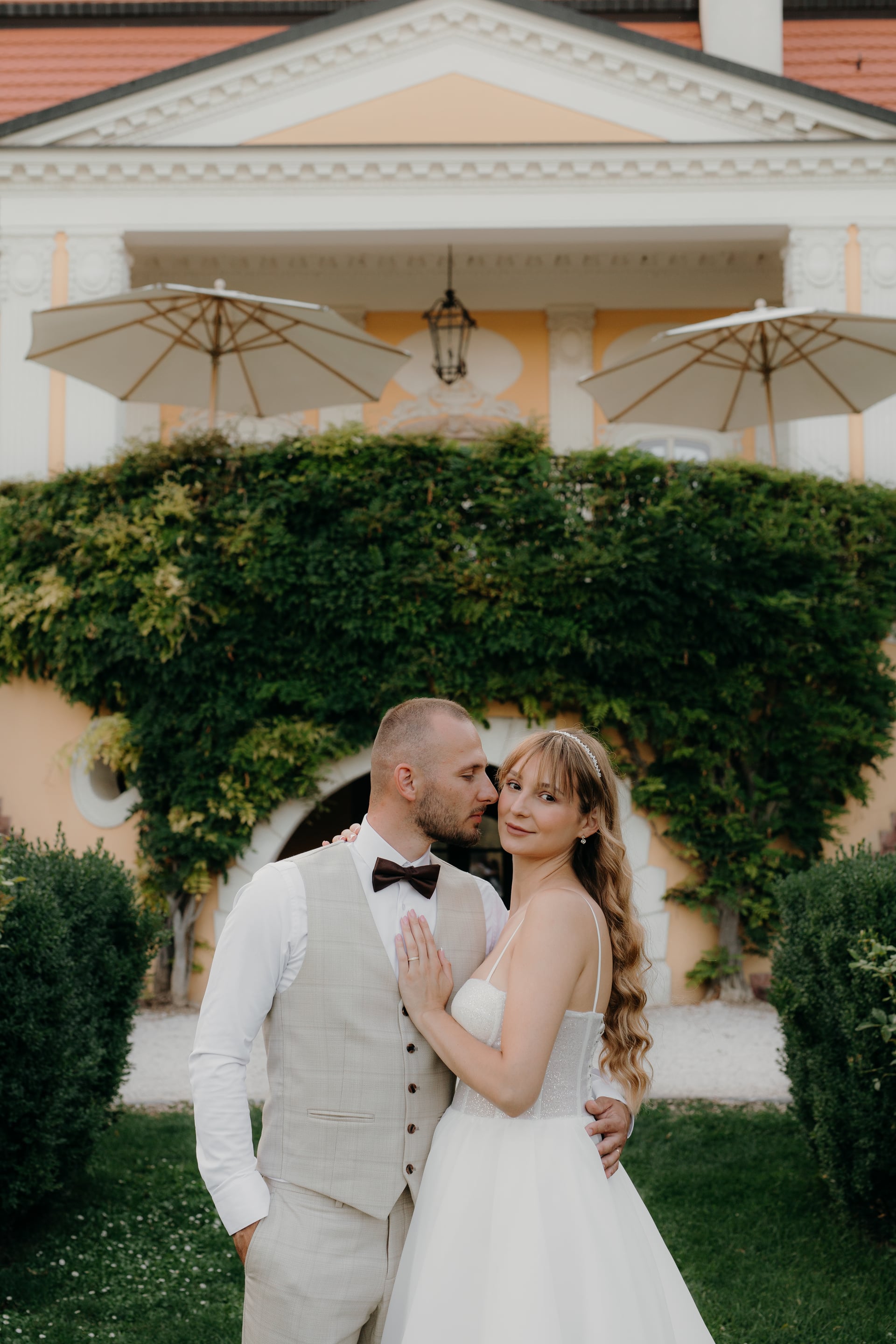 Nevesta v tylových šatách a ženích v béžovom ľanovom vestíku s motýlikom v nežnom objatí pred žltým barokovým kaštieľom so slnečníkmi a brečtanom