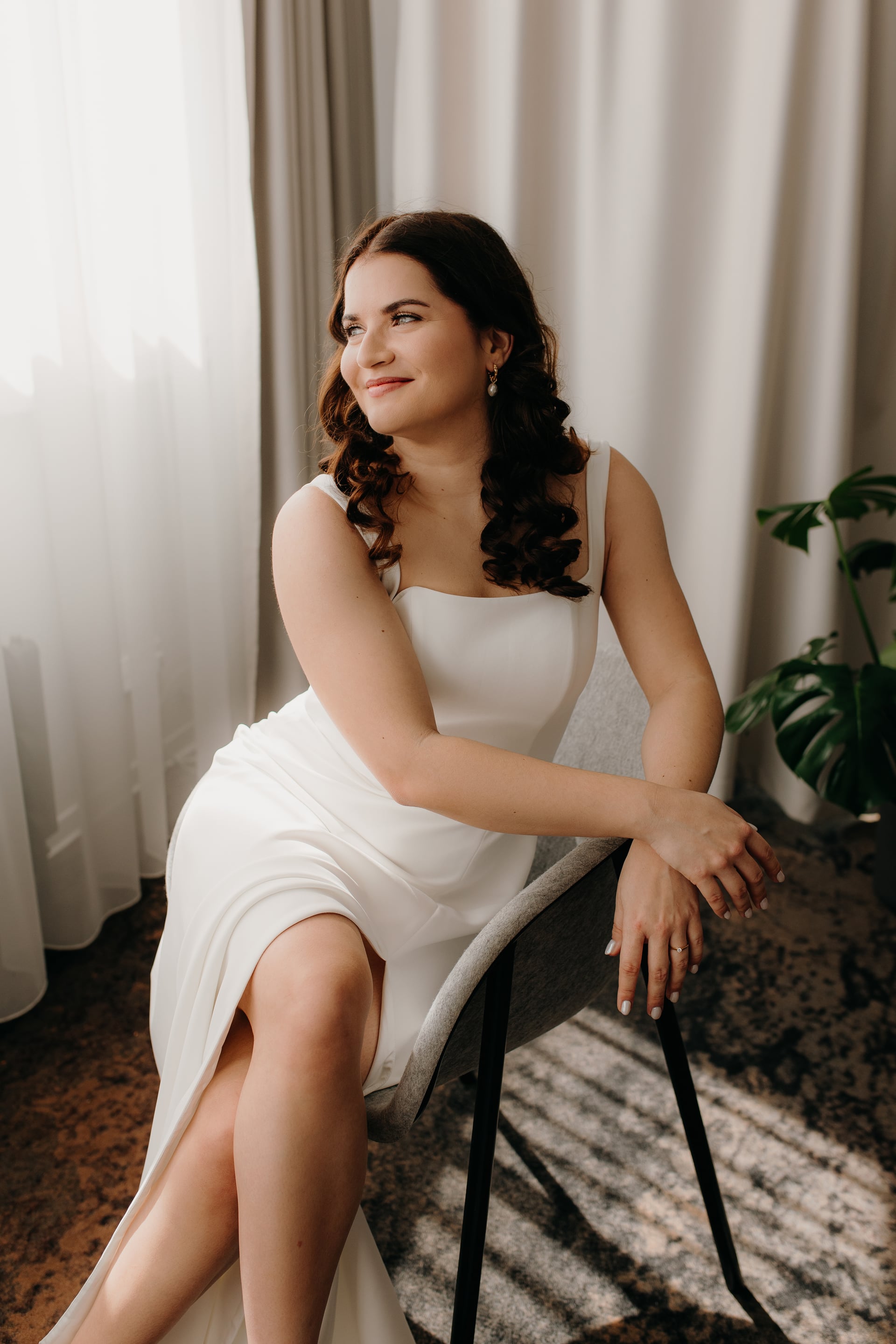 Nevesta v bielych saténových šatách so štvorcovým výstrihom sedí na šedom kresle pri okne s tylovými závesmi — úsmev s pohľadom nabok
