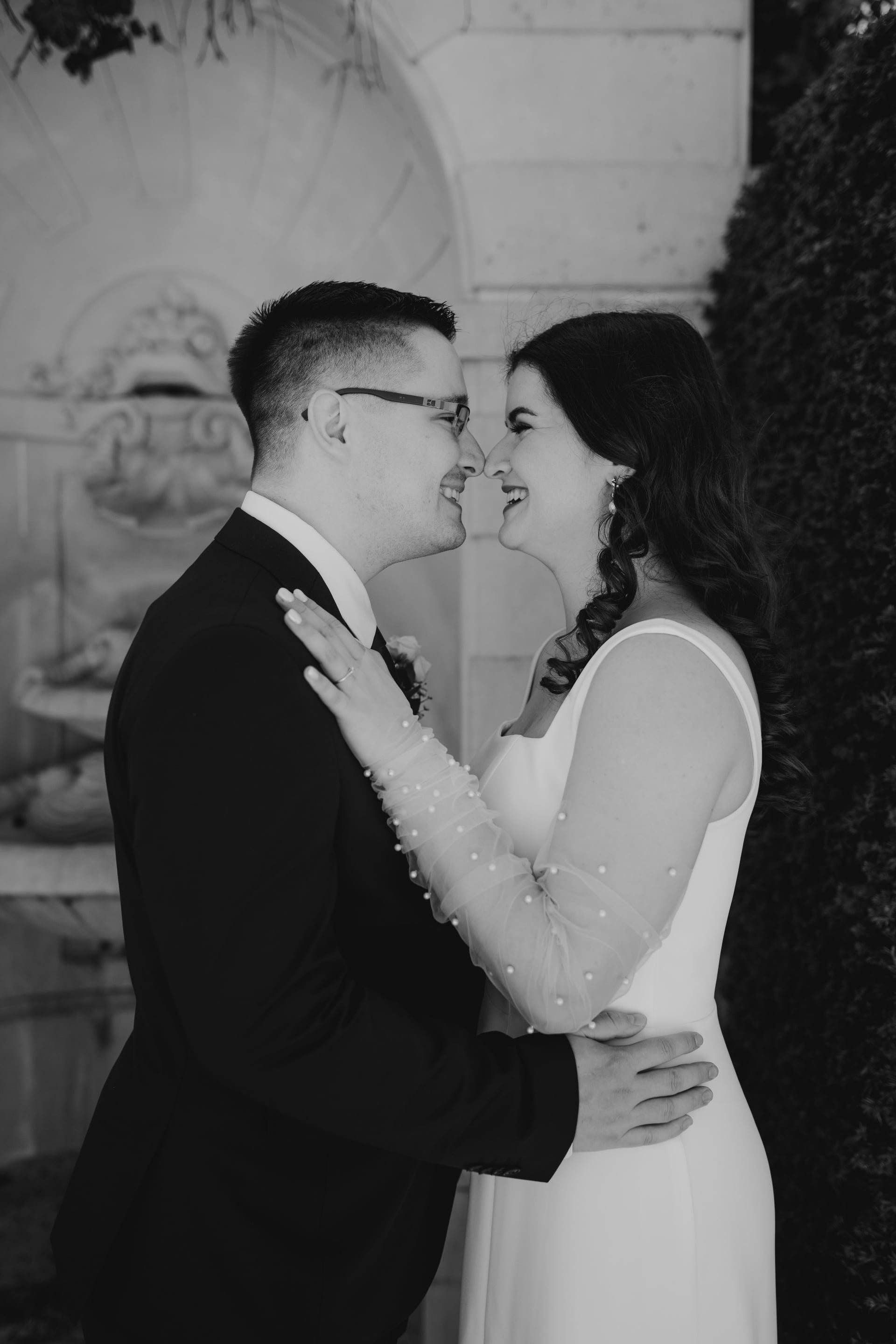 Čiernobiely svadobný portrét — nevesta v šatách s tylovými rukávmi zdobenými perlami a ženích v okuliaroch a tmavom obleku sa smejú nos k nosu pri kamennej stene v záhrade