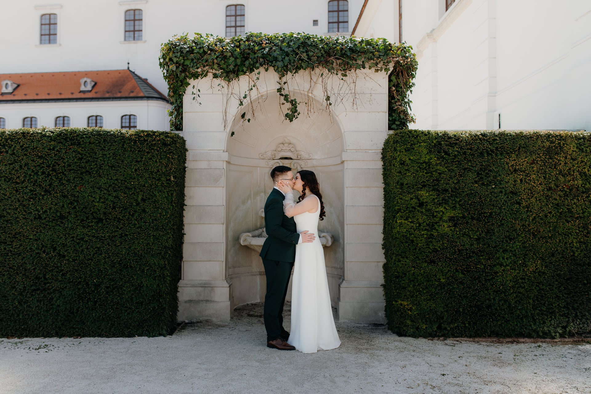 Nevesta a ženích sa bozkávajú pri kamennej fontáne vo formálnej záhrade — strihané živé ploty a baroková budova v pozadí