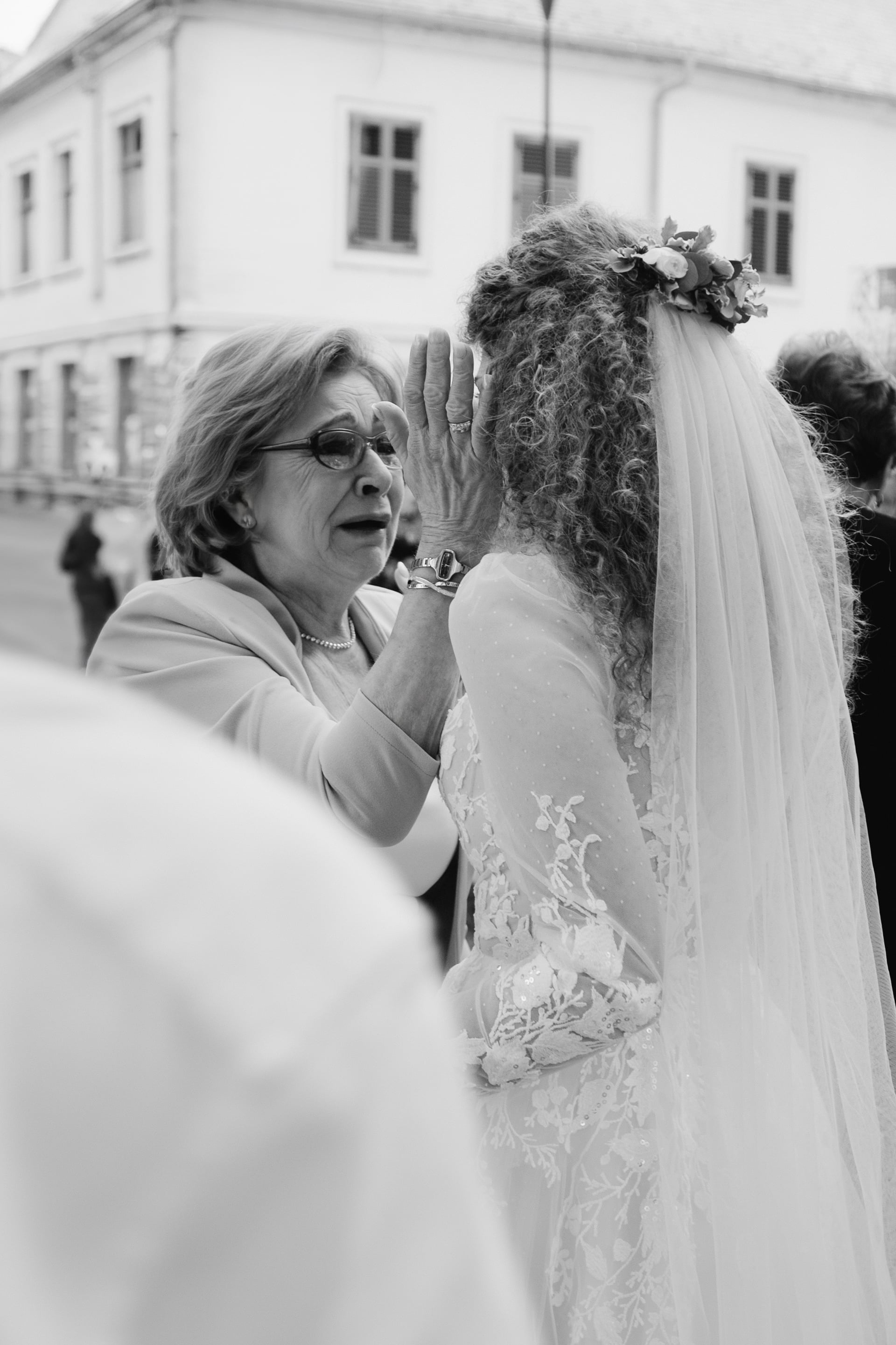 Čiernobiela emotívna scéna — staršia žena s okuliarmi drží v rukách tvár nevesty v čipkovaných šatách so závojom a kvetinovým venčekom na starobylej ulici