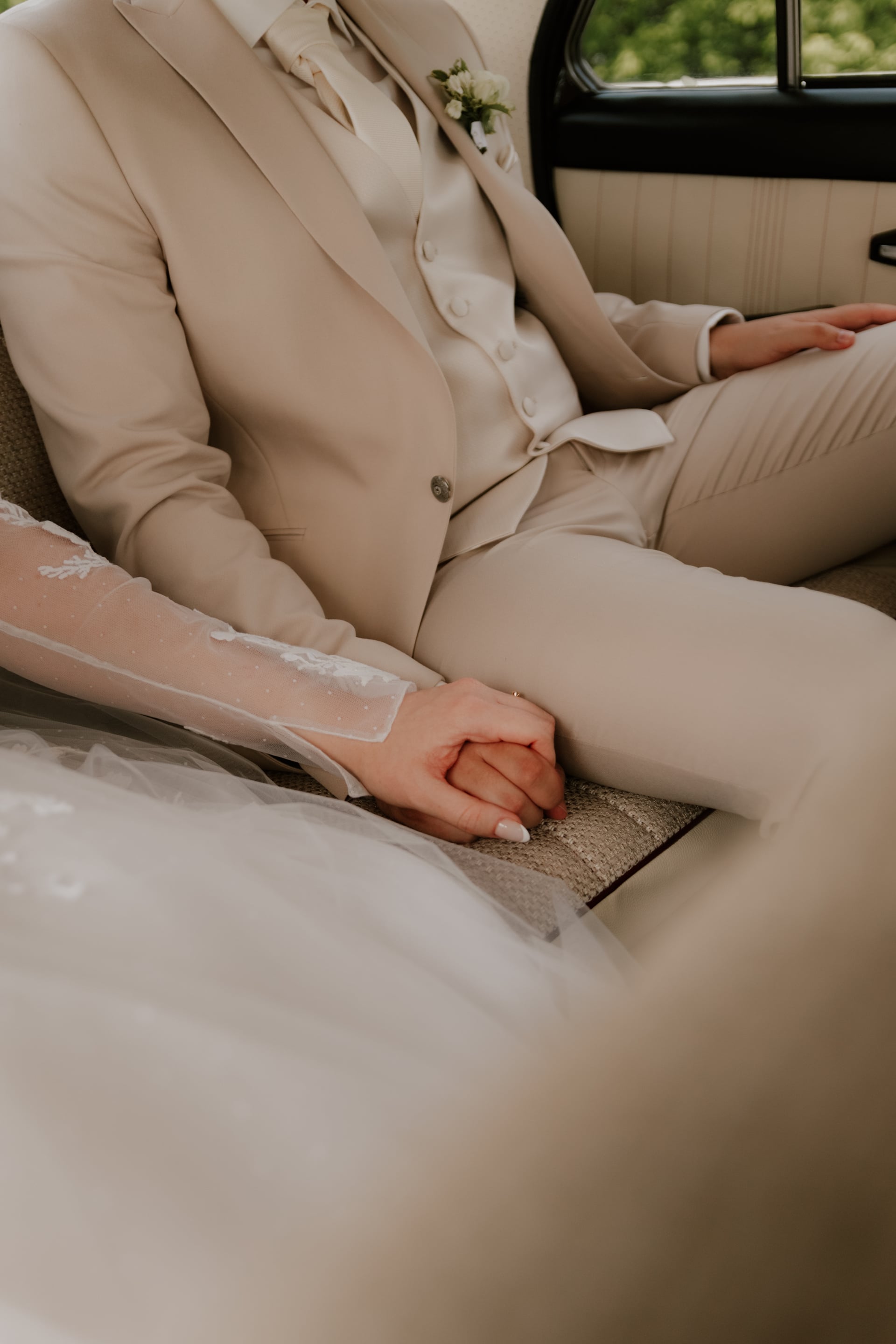 Spojené ruky nevesty v čipkovom rukáve a ženícha v béžovom svadobnom obleku s kravatou na zadnom sedadle klasického auta