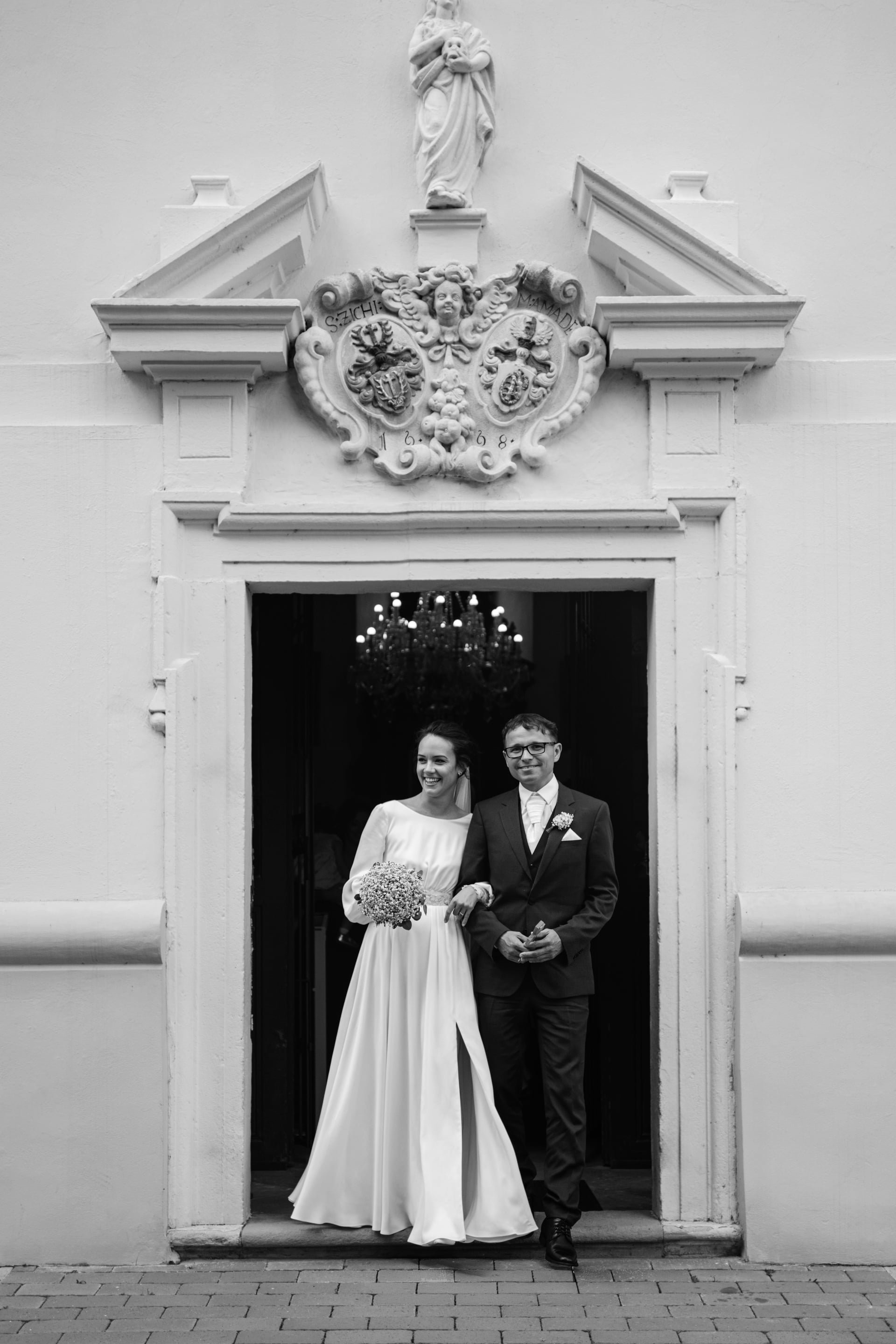 Čiernobiely svadobný portrét — nevesta v hodvábnych šatách s dlhými rukávmi a kyticou a ženích v obleku stoja v portáli barokovej kaplnky so zdobenou kartušou a sochou