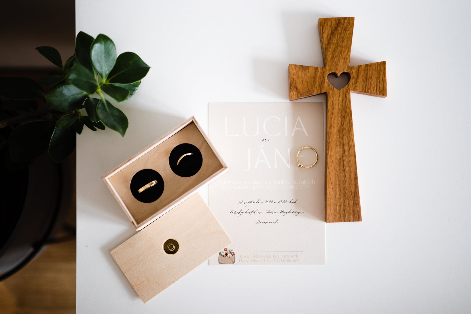 Flat-lay svadobných detailov — dve obrúčky v drevenom puzdre na čiernom zamate, zásnubný prsteň na oznámení, drevený kríž so srdcom a zelená rastlina