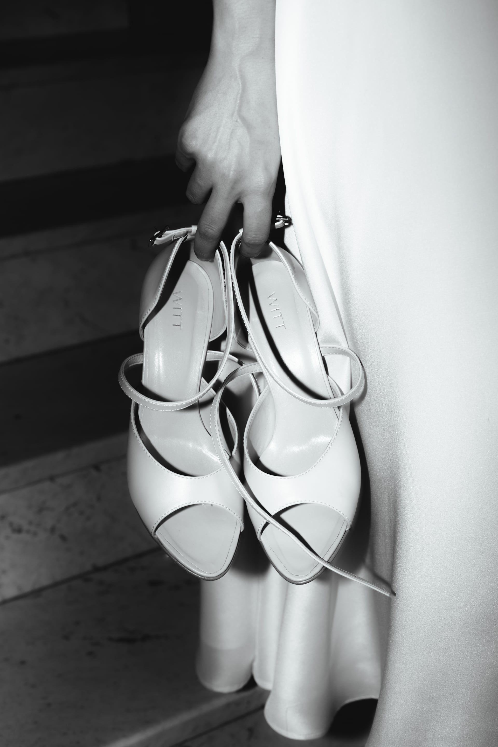 Čiernobiely detail — nevesta drží pár bielych remienkových sandálov za remene pri hodvábnych šatách na kamenných schodoch