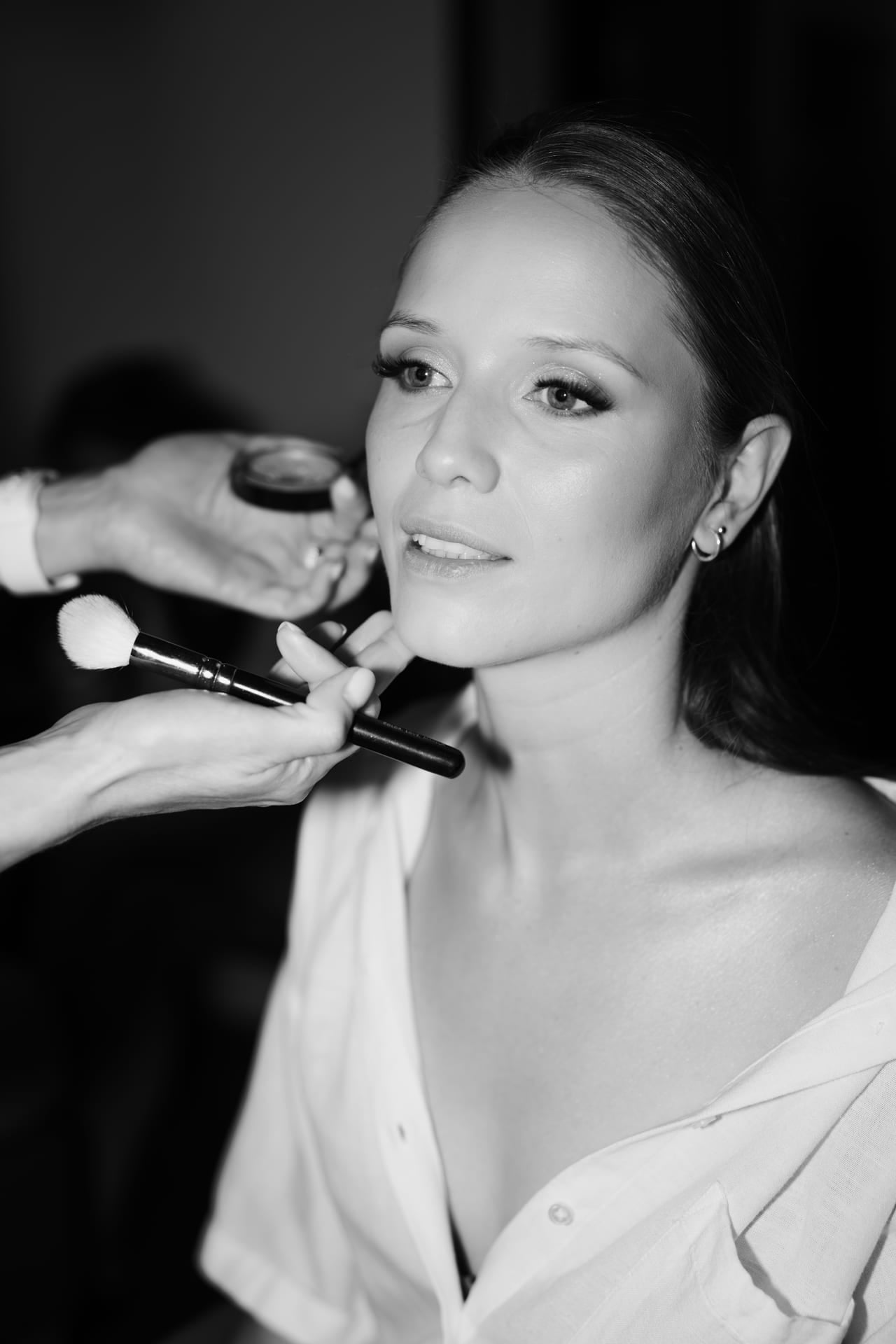 Čiernobiely detail nevesty počas líčenia — ruky vizážistky so štetcom na brade a kompaktom pri tvári, vlasy sčesané dozadu, biela košeľa