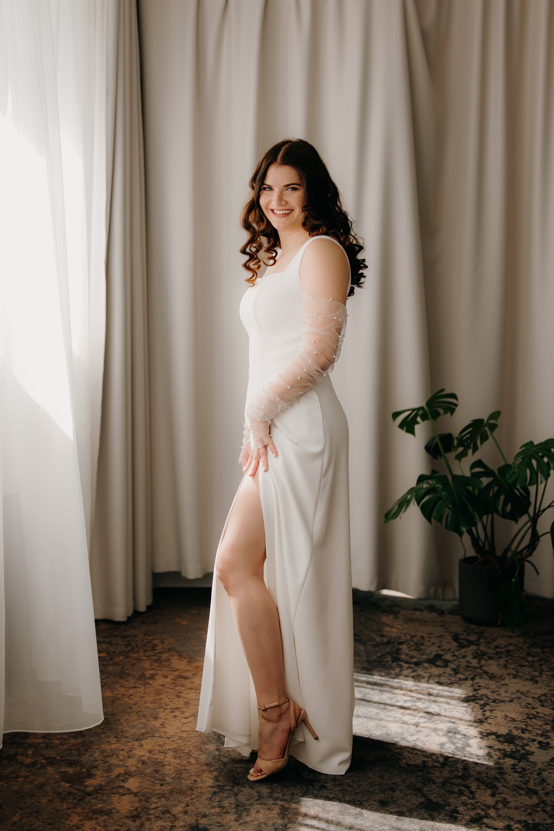 Nevesta stojí v bielych saténových šatách s jedným tylovým rukávom zdobeným perlami a vysokým rozparkom — celkový portrét s úsmevom pri okne