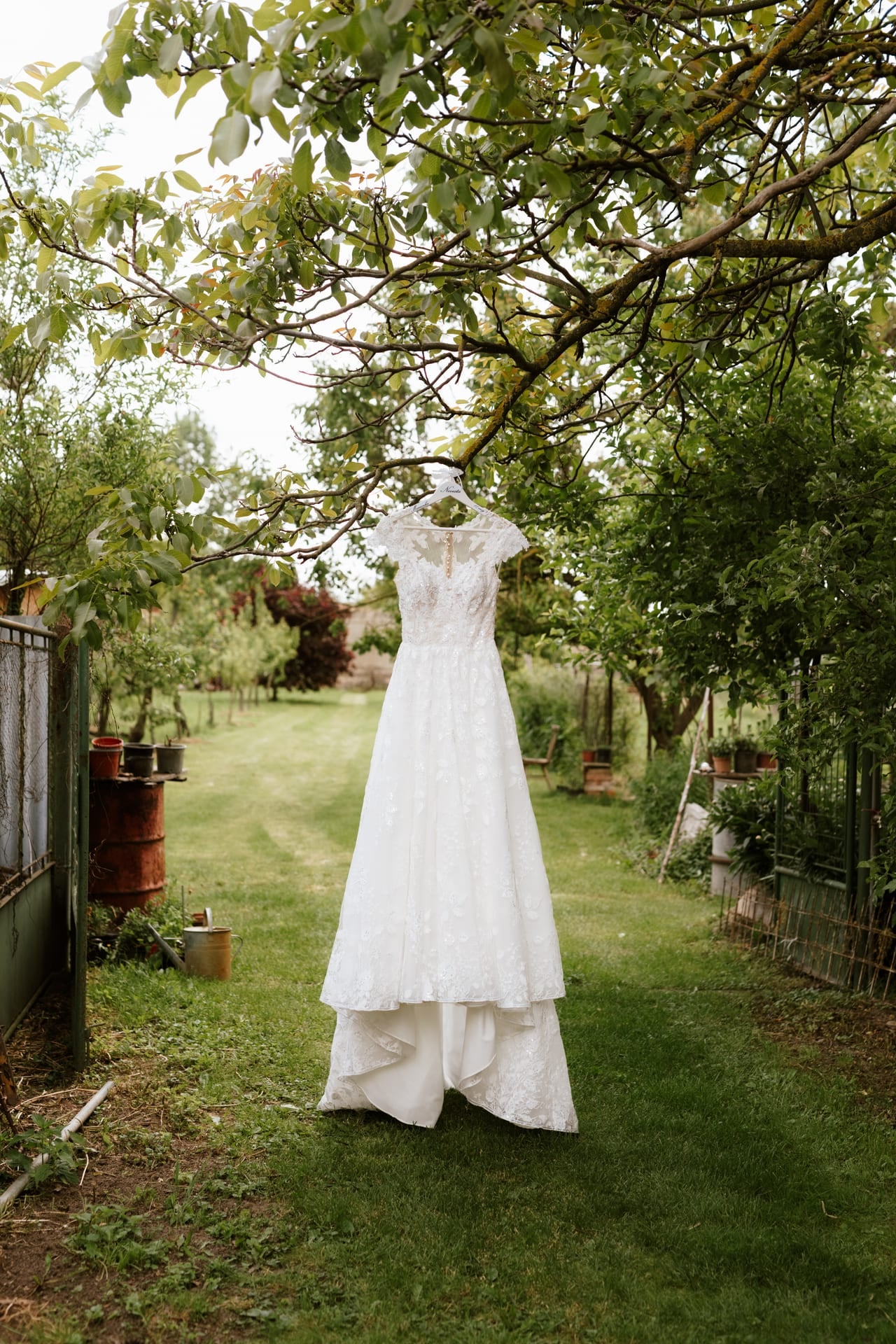 Svadobné šaty s čipkovaným živôtikom a tylovou sukňou visiace na vetve stromu v záhrade — rustikálny kút dvora s drevenou stavbou a kvetinovými kvetináčmi