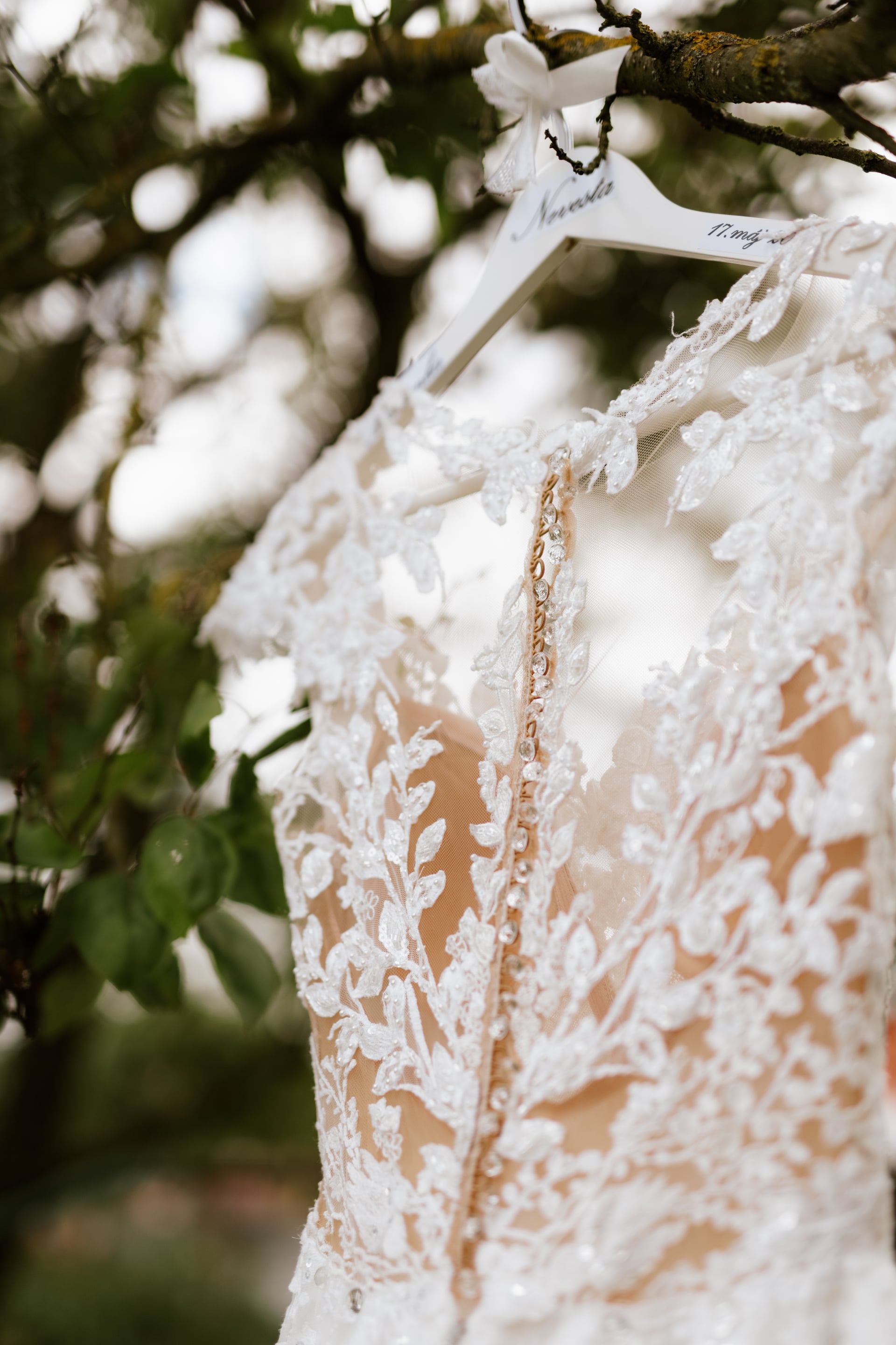 Detail čipkovaných svadobných šiat zavesených na vetve ovocného stromu — drevený vešiak s nápisom Nevesta a zdobený centrálny pás šiat s kamienkami