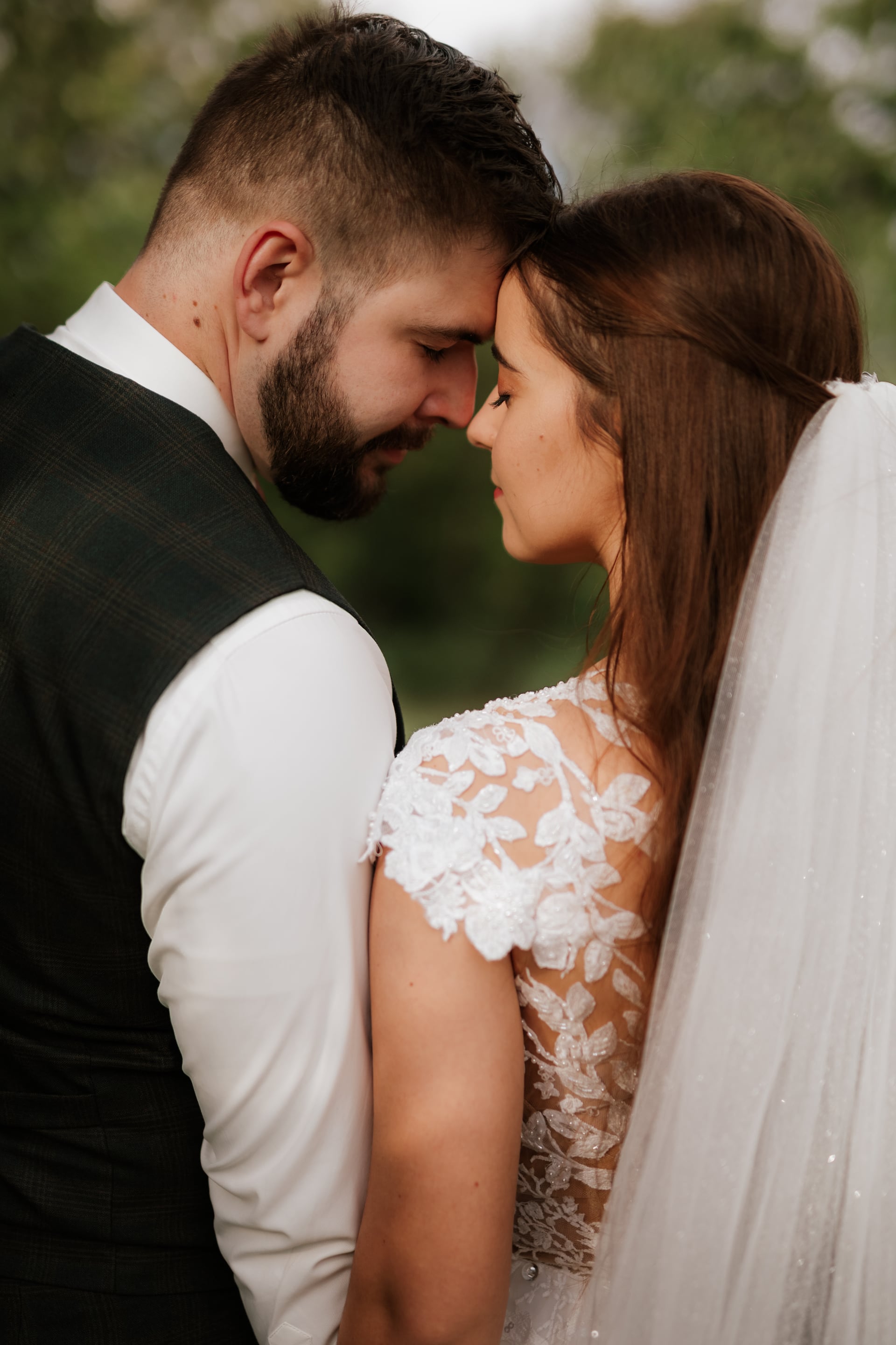 Ženích v károvanej vestíku a bielej košeli a nevesta v čipkovaných šatách so závojom sa dotýkajú čelami so zatvorenými očami — tichý moment v záhrade