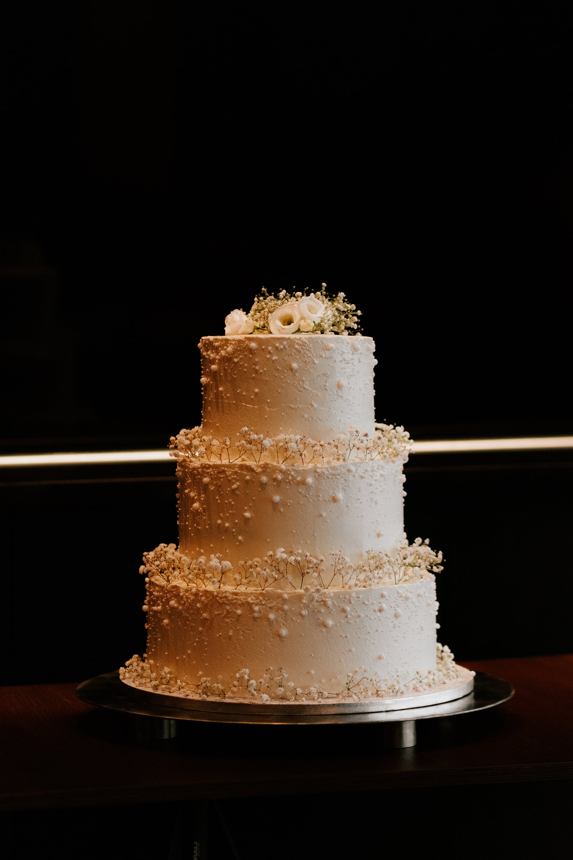 Trojposchodová biela svadobná torta s textúrou a perličkami, zdobená gypsomilou a drobnými bielymi ružami na kovovom tortovom podnose v tmavom priestore