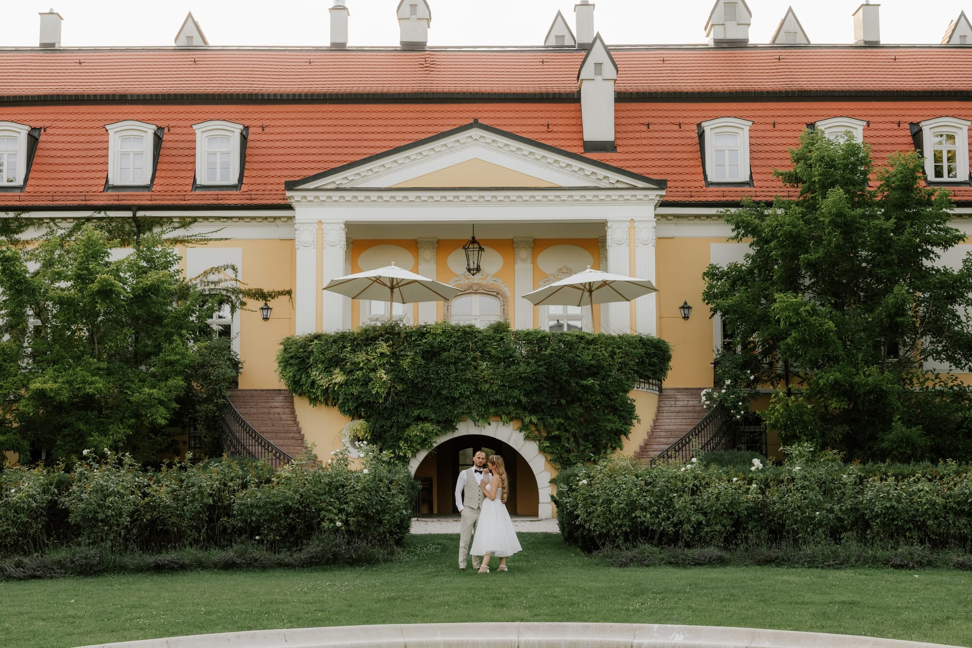Nevesta a ženích v objatí pred žltým klasicistickým kaštieľom — trávnatá záhrada so strihanými živými plotmi a slnečníkmi pri vchode