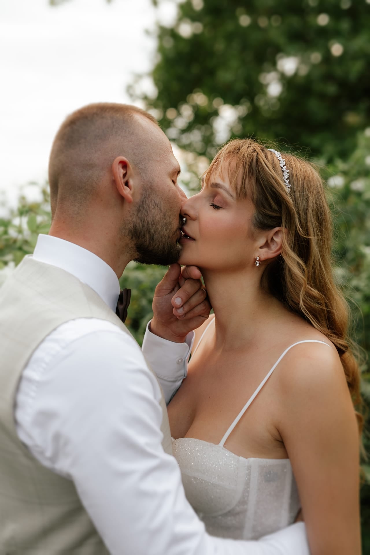 Detailný svadobný portrét — ženích v béžovom vestíku s motýlikom jemne pridržiava nevestinu tvár a bozkáva ju do líca, nevesta s trblietavým živôtikom a zdobenou čelenkou v záhrade