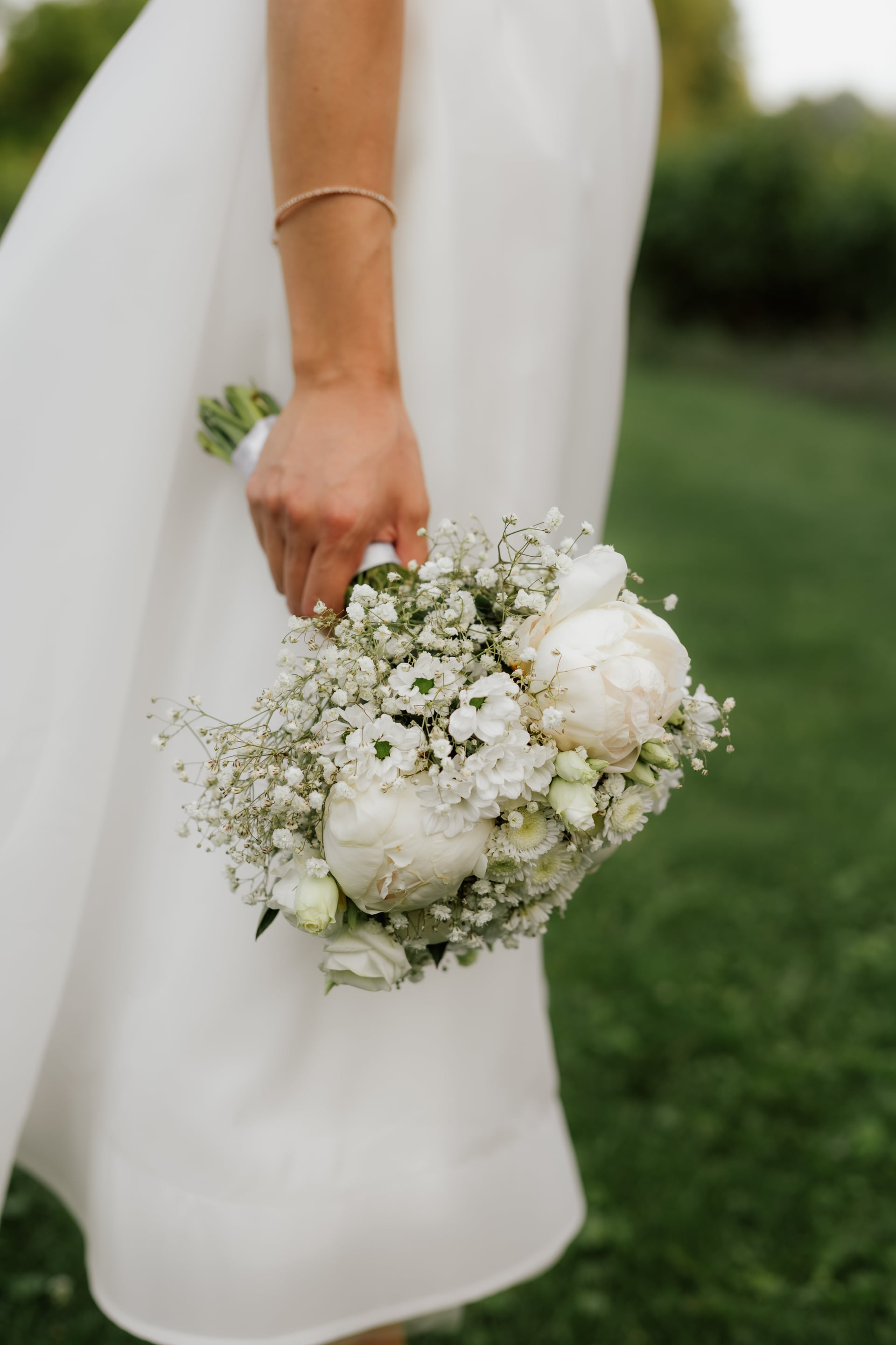 Nevesta v bielych saténových šatách drží svadobnú kyticu z bielych pivónií, lisiantusov a gypsomilky — tenký zlatý náramok na ruke, zelená záhrada v pozadí