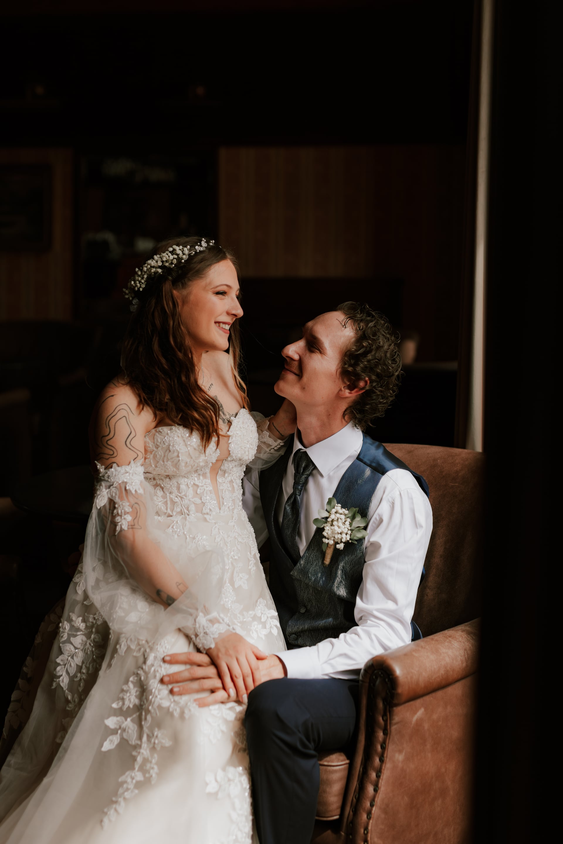 Nevesta v čipkovaných šatách s puffy tylovými rukávmi a kvetinovým venčekom z gypsomily a ženích s petrolejovou jacquardovou vestou sa na seba usmievajú v koženom klubovom kresle